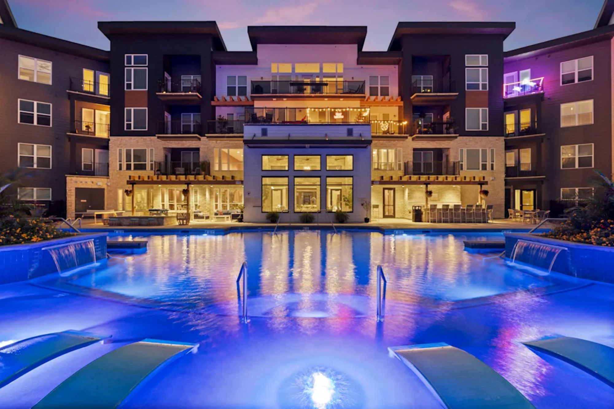 An illuminated apartment swimming pool glowing with blue lighting at dusk in front of a multi-story clubhouse.